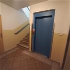 Vstupní hala bytového domu s dlažbou, modrým výtahem a schodištěm, čisté stěny a moderní design pro pohodlný přístup.